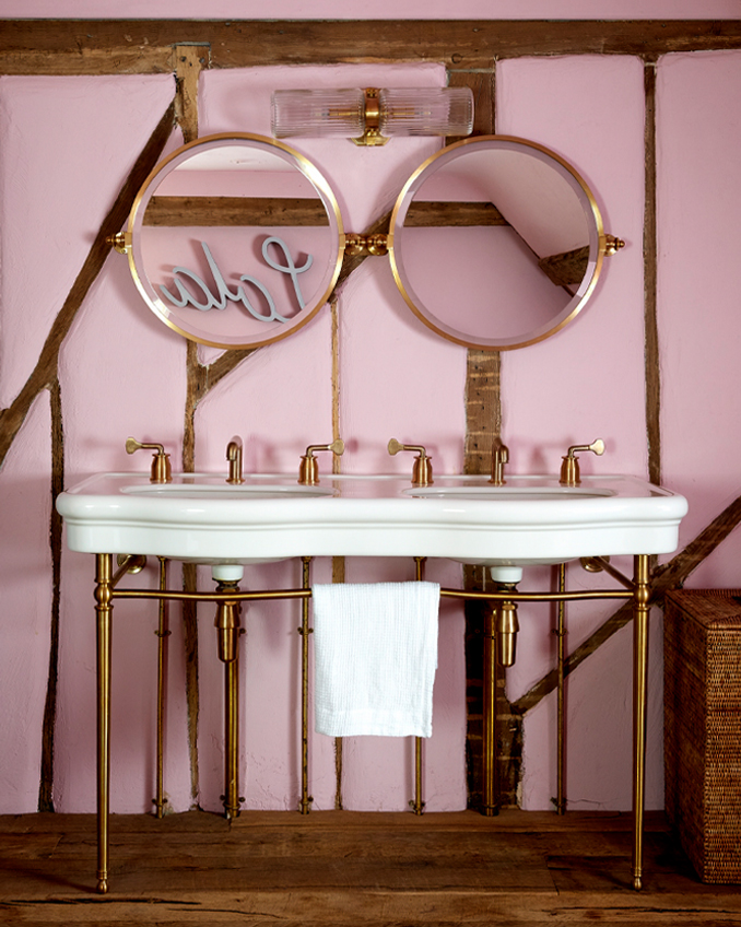 A traditional white bathroom basin in a bathroom with pink walls. The double mirror and the open pipes have a brass finish from Ana Engelhorn, interior designer Surrey.