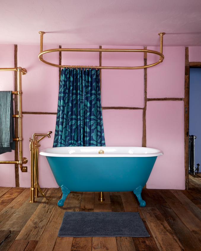 A beautiful blue, free-standing, roll-top bath in a pink bathroom with copper pipes and plumbing on show. The room is finished off with a blue shower curtain from Annika Reed.