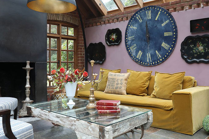 Luxuriously designed living room interior with vaulted ceiling, mustard-coloured couch, upholstered armchairs, fireplace and statement pieces including an oversized wall clock as a focal point above the couch.
