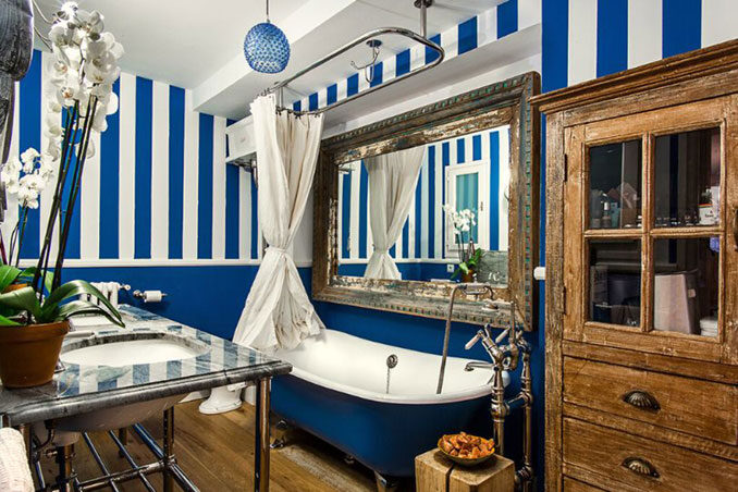 Eclectic interior design luxury bathroom decor with blue and white striped walls, ball and claw bathtub, wooden flooring and cabinetry in a stylish Barcelona apartment designed by Ana Engelhorn.